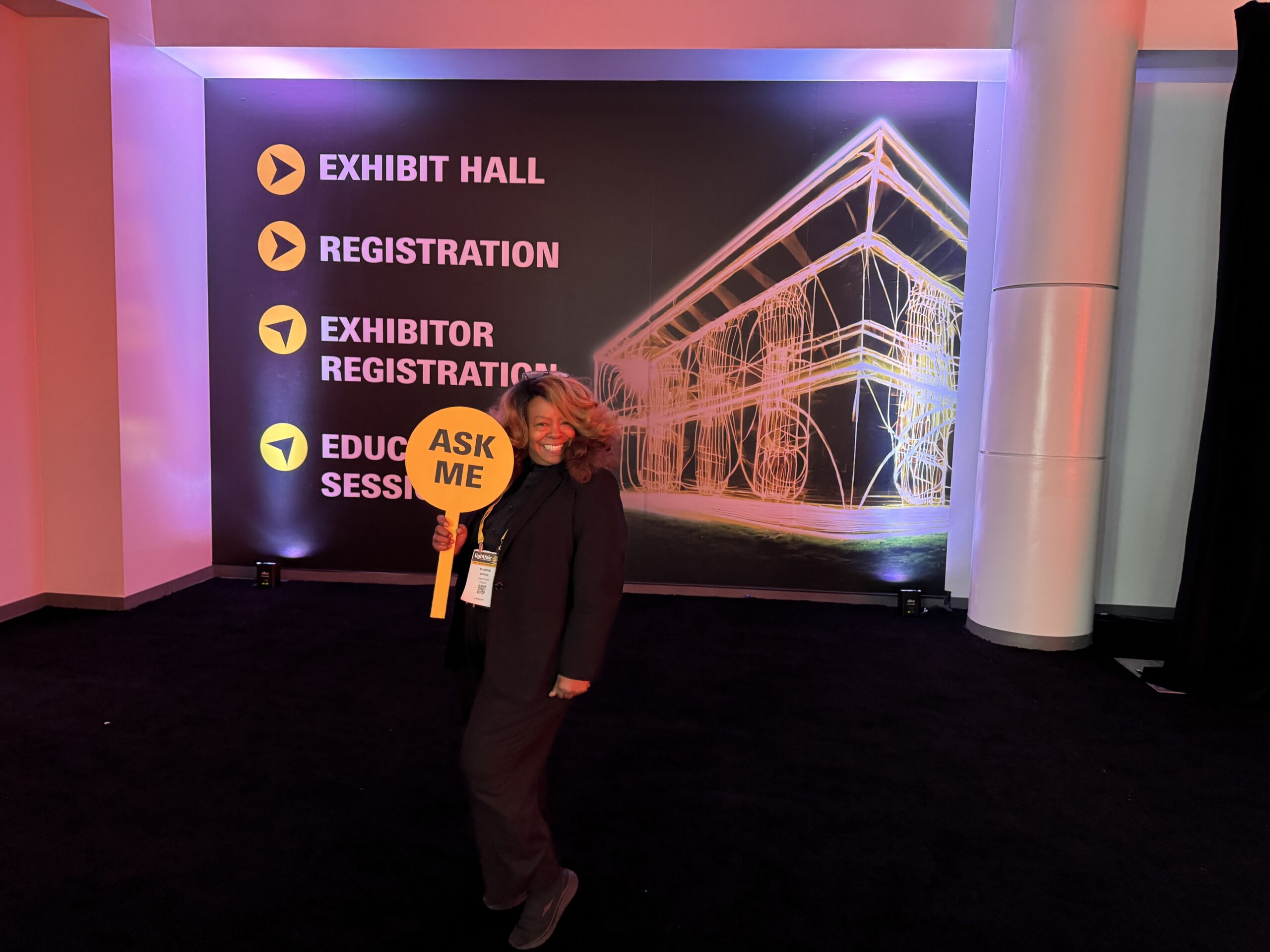 Lady holding up sign ASK ME. LightFair has plenty of people